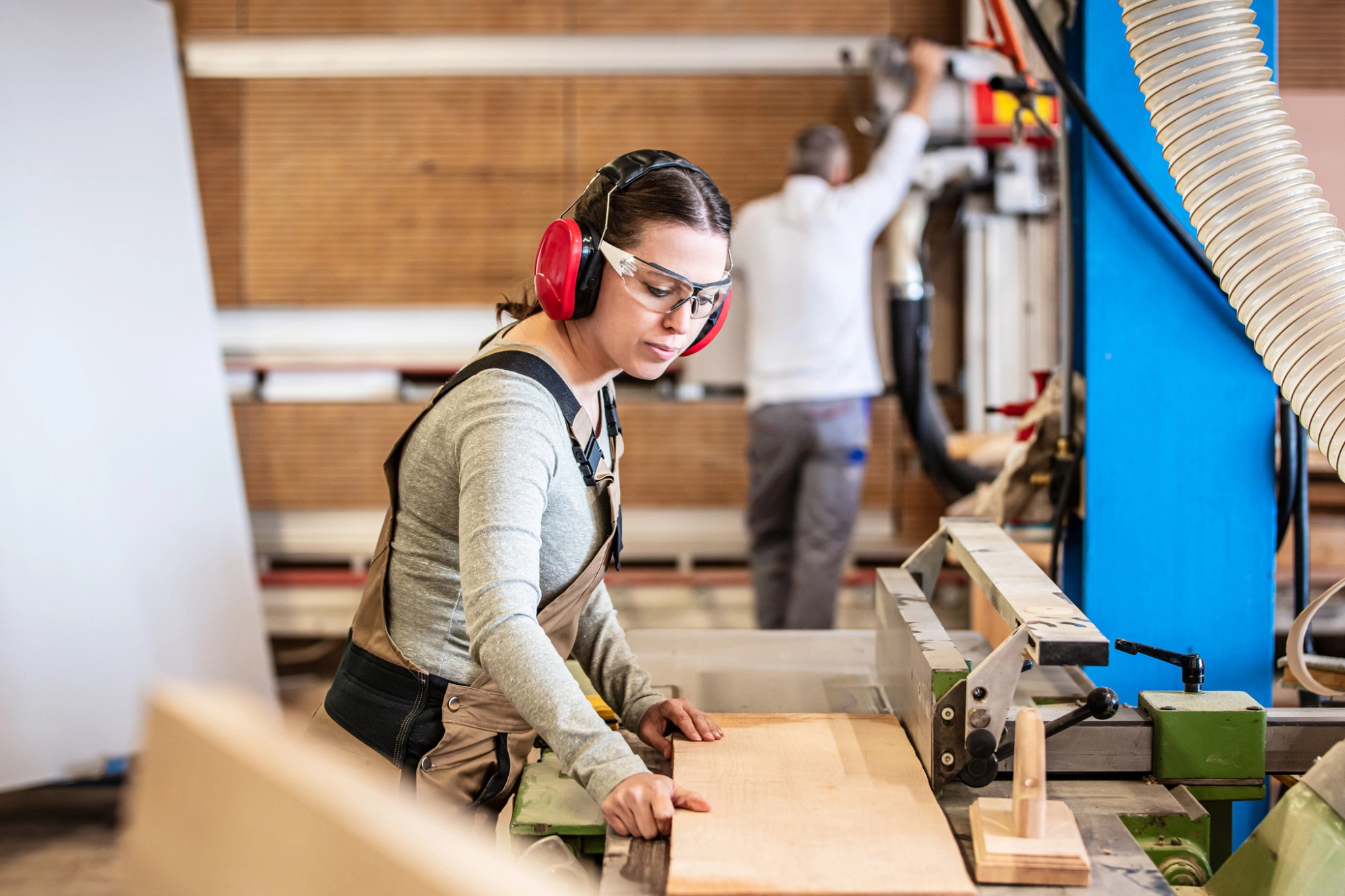 Eine Schreinerin mit Gehörschutz und Schutzbrille arbeitet in einer Werkstatt mit Holz.
